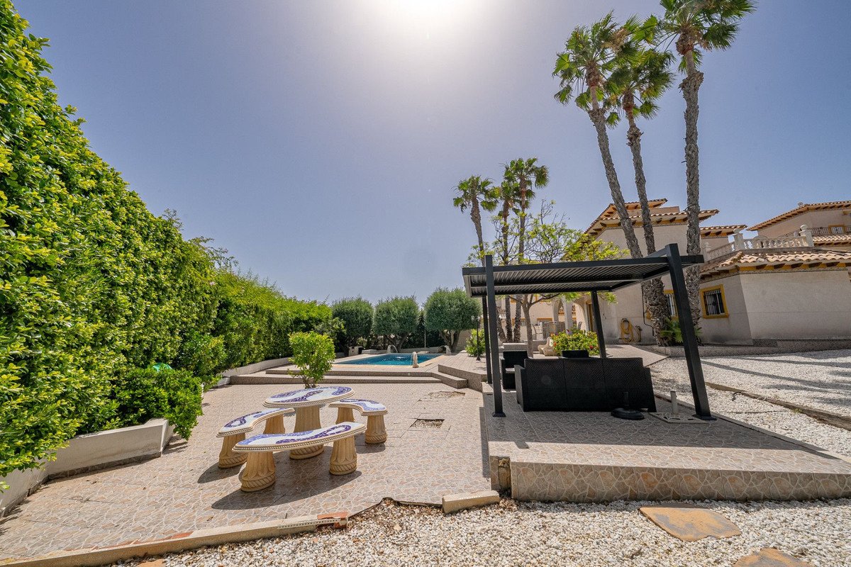Outdoor area with a swimming pool, shaded seating area, picnic table, palm trees, and lush greenery.