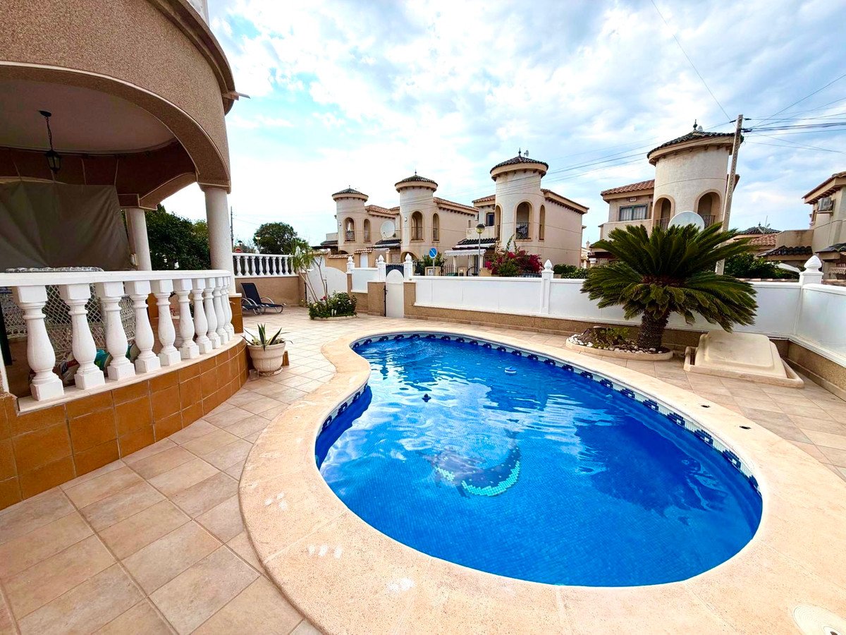 A private swimming pool with blue water and a tiled patio area, with houses and a palm tree in the background.