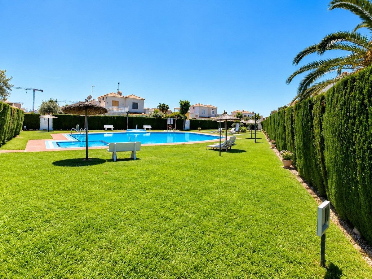 Outdoor communal swimming pool with lounge chairs and umbrellas on a sunny day, surrounded by green grass and hedges.