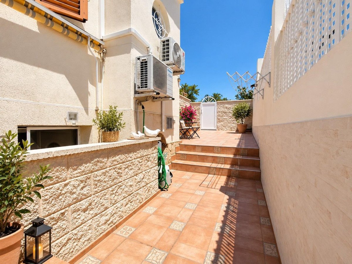 Sunny outdoor terrace with tiled steps leading up to a white door, potted plants, and air conditioning units on the building wall.