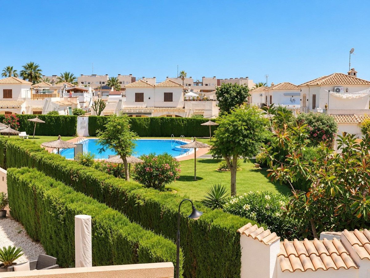 View of a villa complex with a swimming pool, lush gardens, and trees under a clear blue sky.