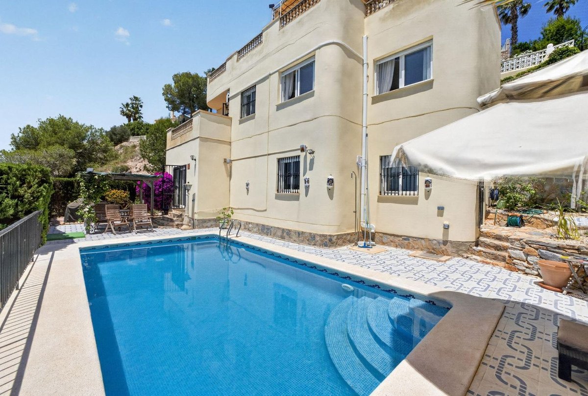 A spacious villa with a large blue swimming pool and a tiled patio area surrounded by greenery and trees.