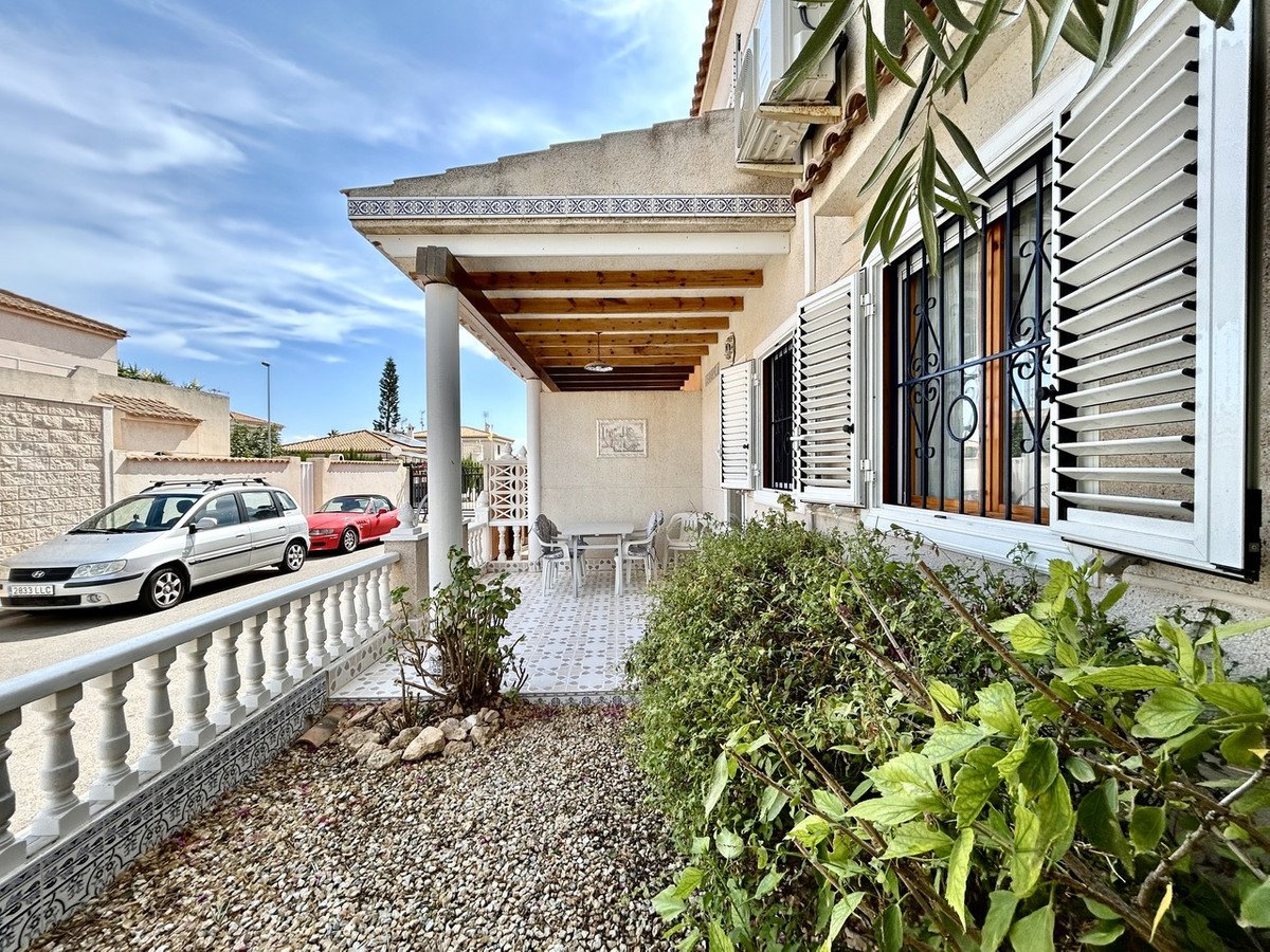 Covered terrace with tiled floor, outdoor furniture, and cars parked outside the property. Lush green bushes in the foreground.