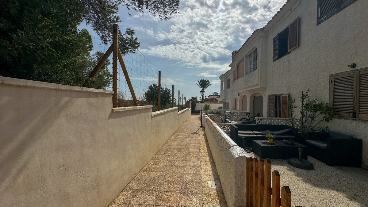 A paved walkway leads towards a terrace with black outdoor furniture, near a white building and lush trees under a cloudy sky.
