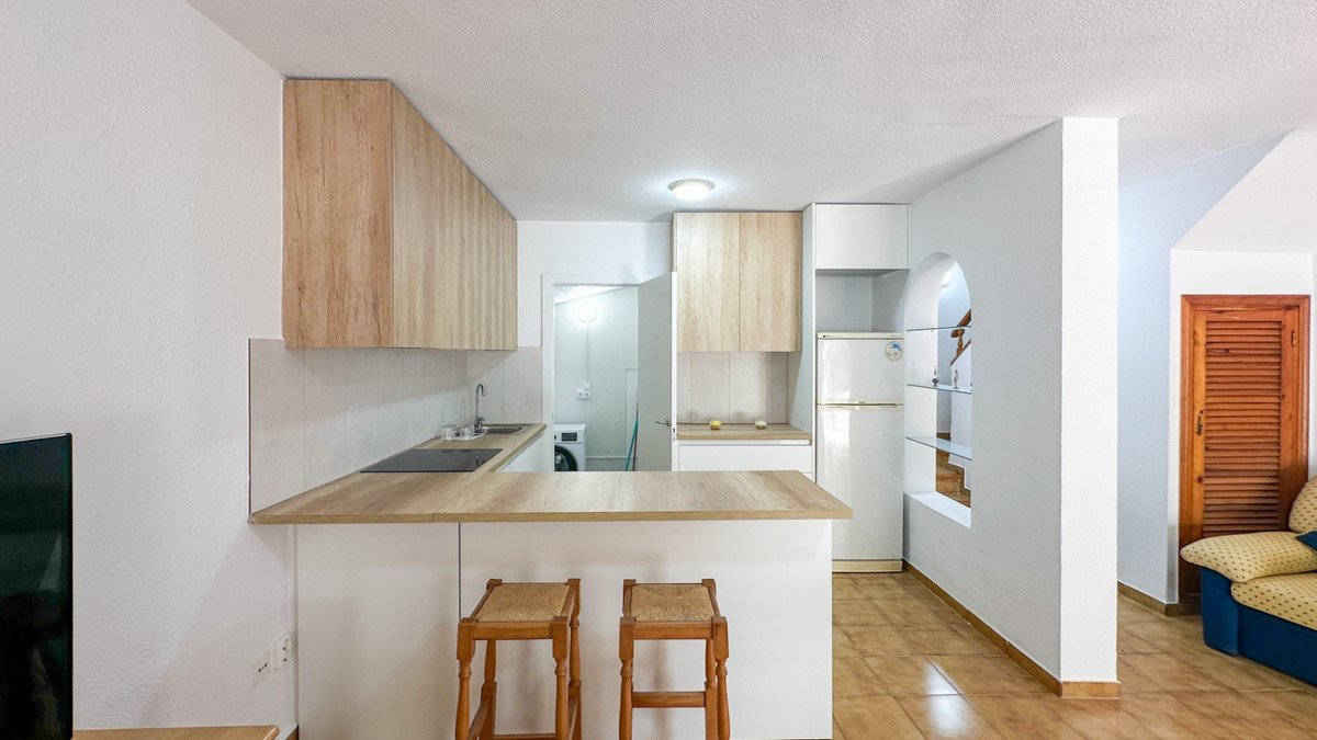 A modern kitchen with wooden cabinets, a breakfast bar with stools, and appliances like a refrigerator and washing machine.