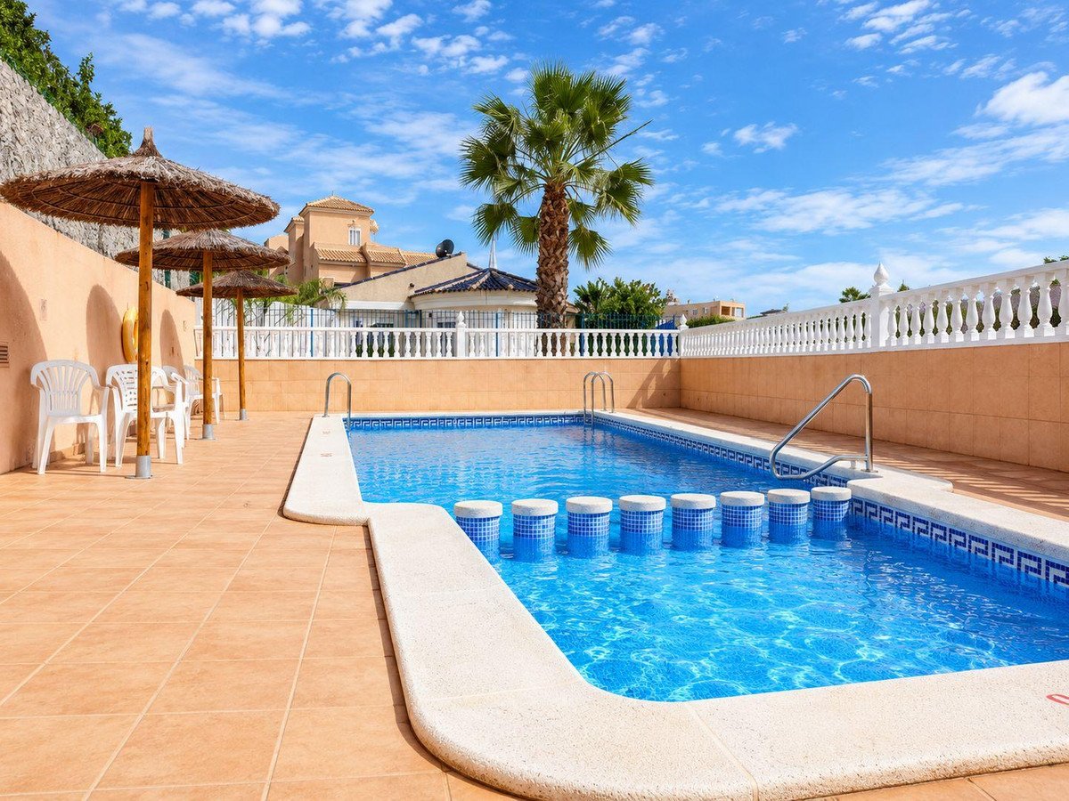 Outdoor swimming pool with blue water, palm tree, and patio seating under straw umbrellas.