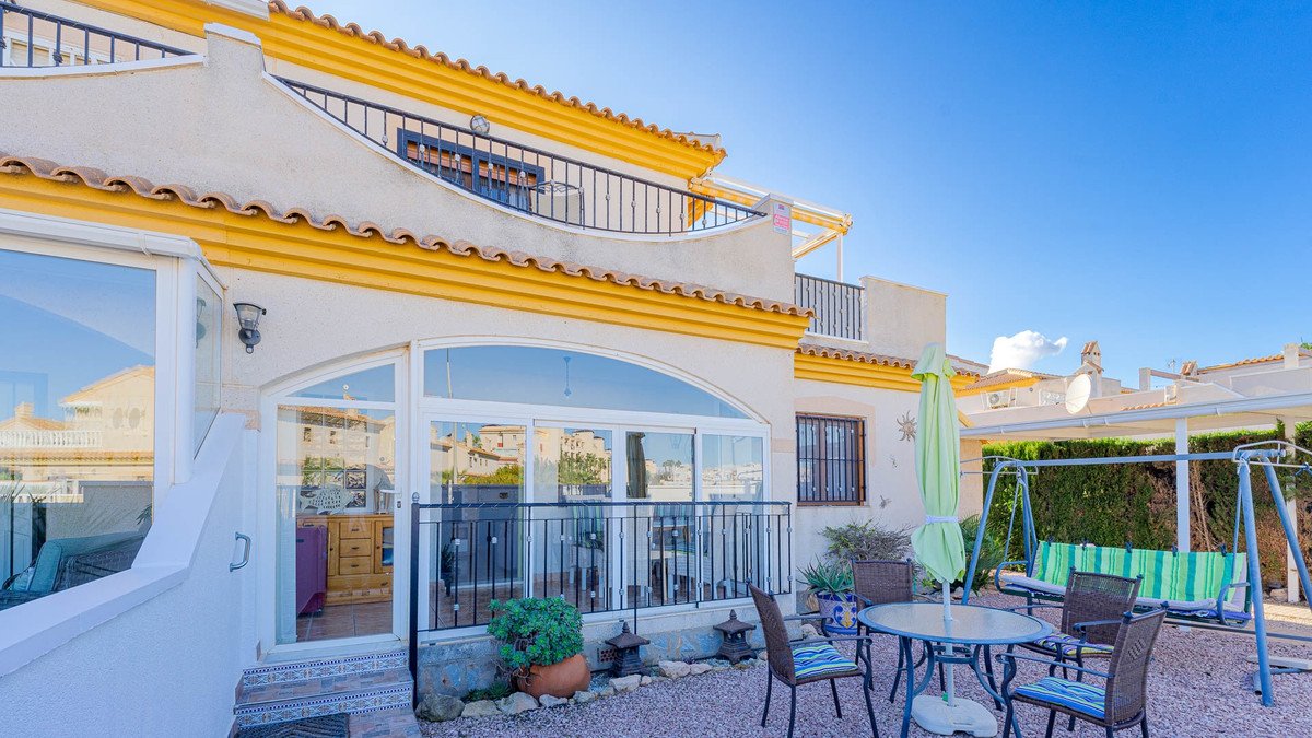 Villa with yellow trim, patio, table, chairs, swing, and garden view under clear blue sky.