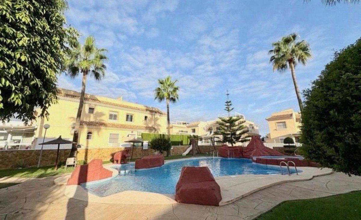 A blue swimming pool with palm trees and resort buildings under a cloudy sky.
