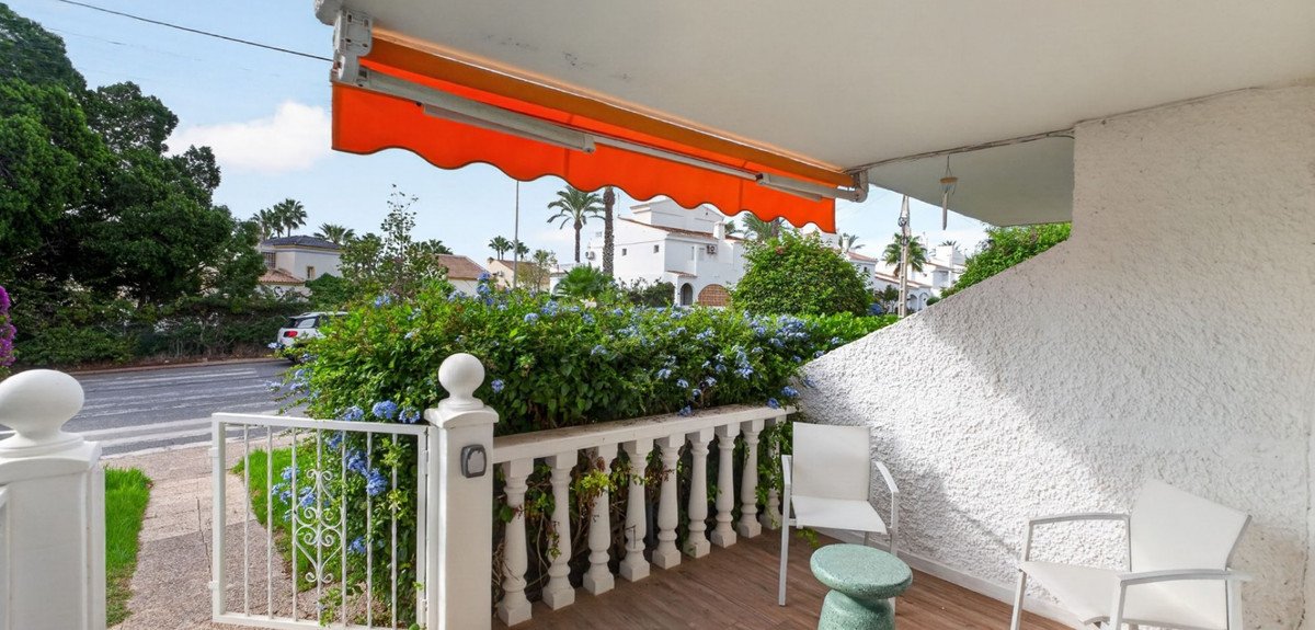 A covered terrace with white chairs, a green table, lush greenery, and an orange awning. Sunny day with palm trees and houses in the background.
