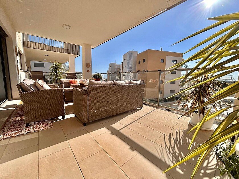 A spacious terrace with wicker furniture, a coffee table, and a glass railing overlooking modern buildings under a sunny sky.