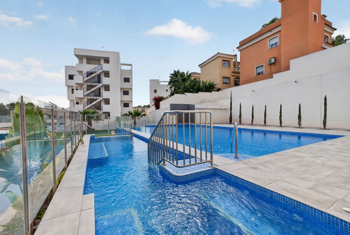A view of a modern outdoor pool complex with blue water and tiled edges, surrounded by contemporary buildings under a blue sky.