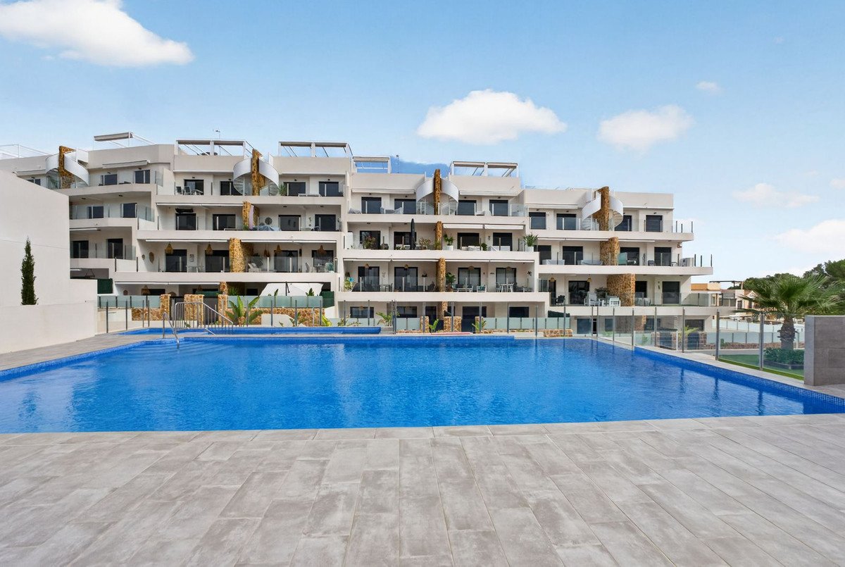 Modern apartment building with balconies overlooking a large blue swimming pool under a partly cloudy sky.