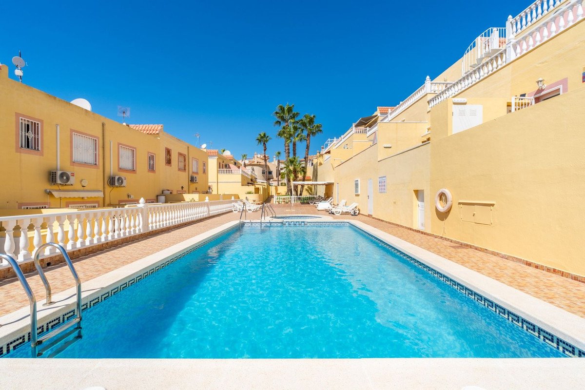 A bright blue swimming pool with palm trees and yellow buildings under a clear blue sky.