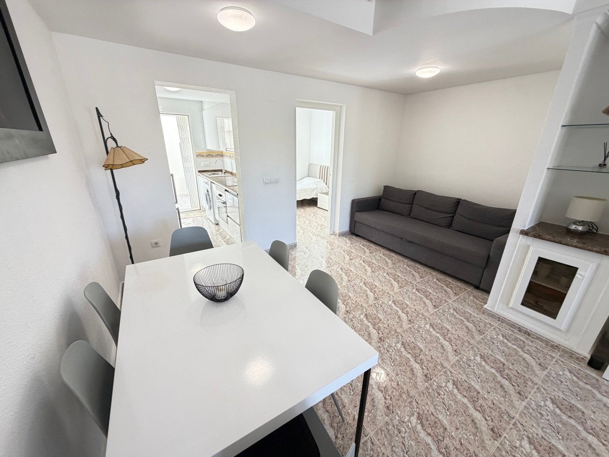 A bright living room with a sofa, dining table, and a glimpse of the kitchenette and bedroom.