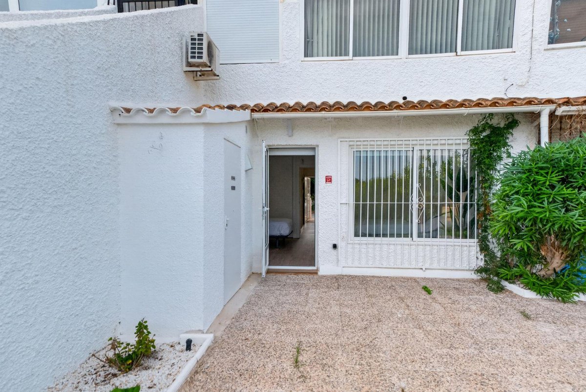 Exterior view of a property showing a gravel terrace, an open door revealing a bedroom, and a window with security bars.
