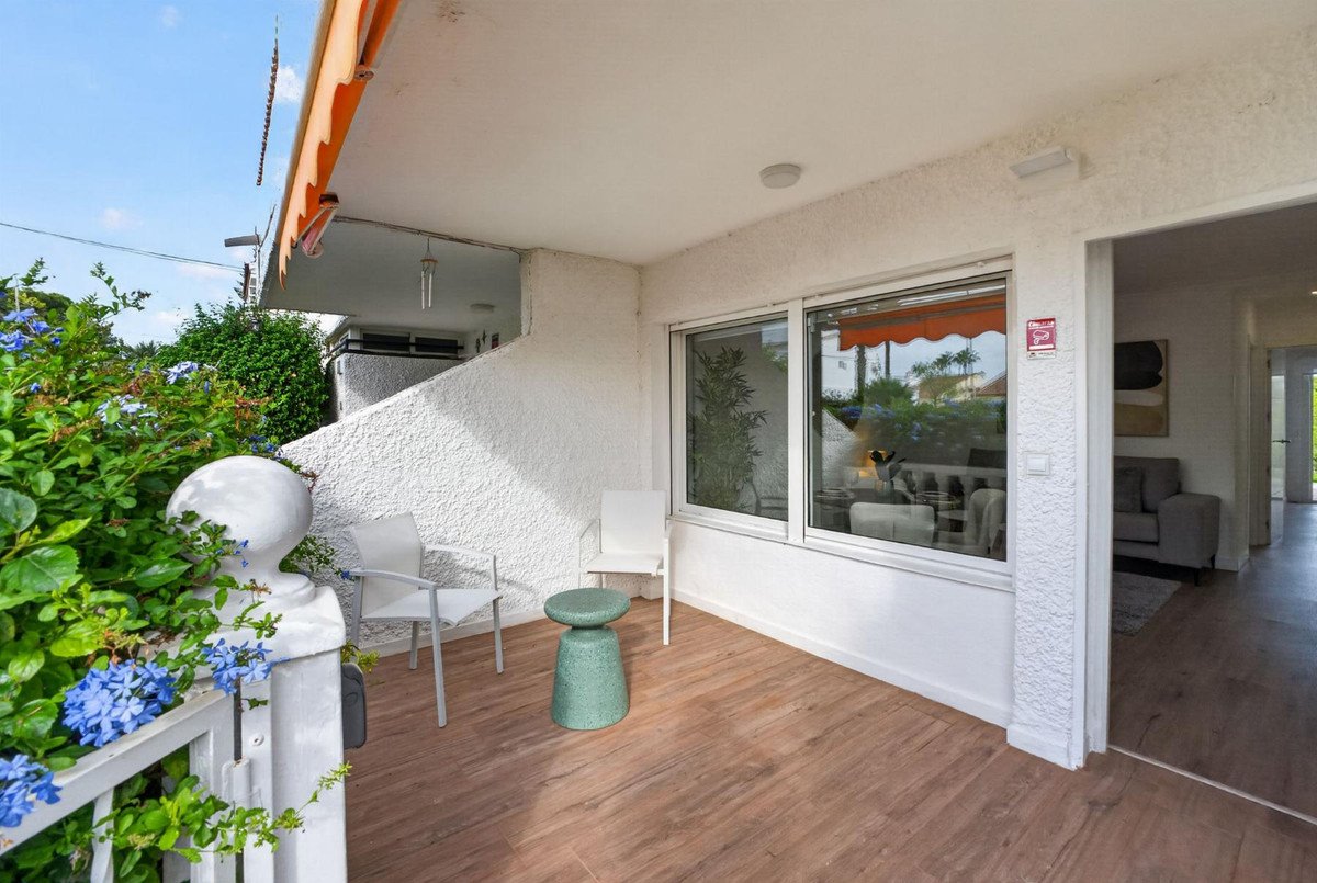 Outdoor terrace with two chairs, a green stool, and lush plants, featuring a large window view of a living room.