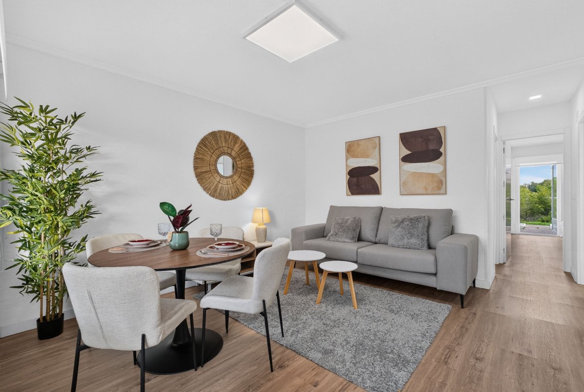 Modern living and dining area with a grey sofa, round dining table, and view of greenery through an open doorway.