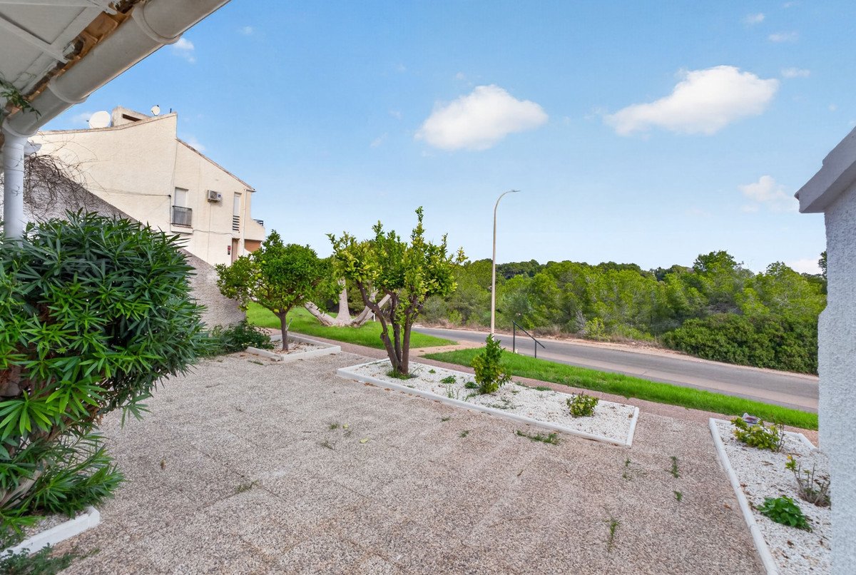 Outdoor patio area with small trees and gravel beds, overlooking a road and lush green trees under a blue sky.