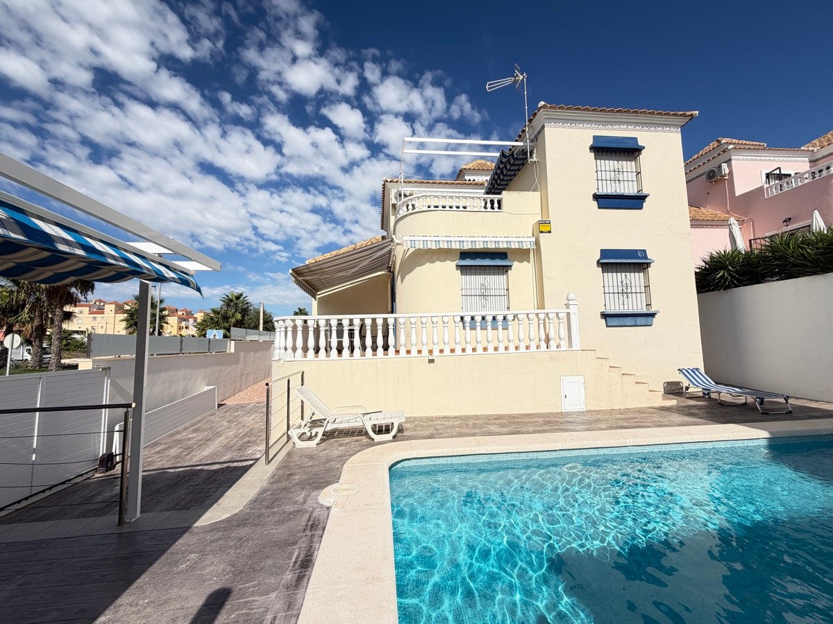 A villa with a swimming pool, terrace, and lounge chairs under a blue sky with clouds.