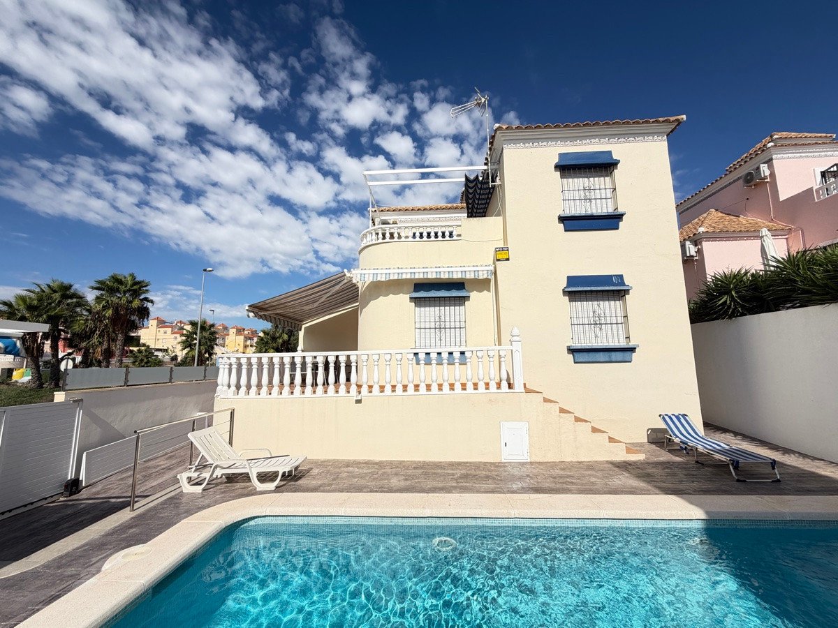 A pale yellow villa with a blue-trimmed balcony, a swimming pool, sun loungers, and palm trees under a cloudy sky.