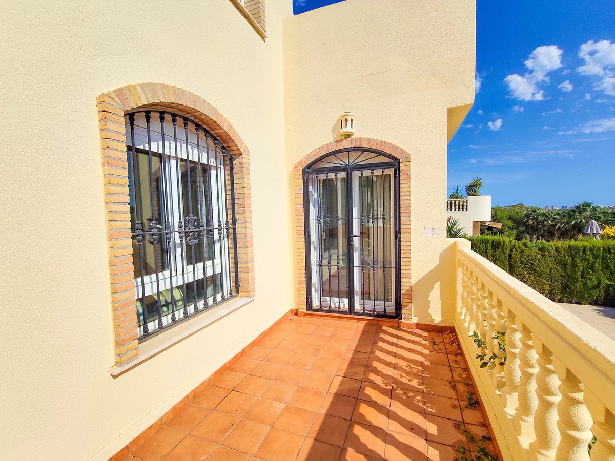 A sunny balcony with terracotta tiles and a view of green hedges under a bright blue sky with clouds.