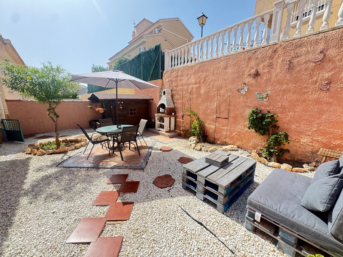 A sunny garden terrace with outdoor dining set, barbecue grill, lounge seating made of pallets, and a small wooden shed.