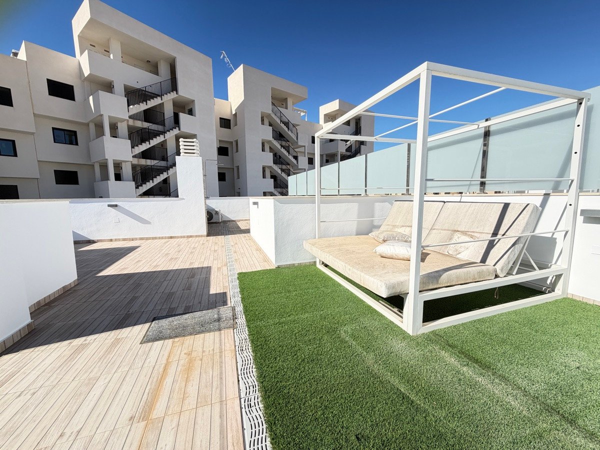 Modern terrace with artificial grass, wooden decking, and a covered outdoor daybed overlooking a contemporary apartment building.
