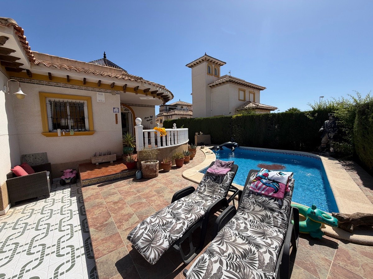 A sunny villa exterior with a private swimming pool, two sun loungers, and garden furniture.