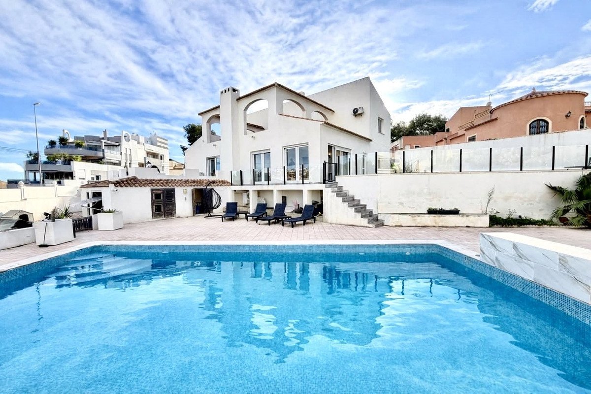 Modern white villa with a large swimming pool, lounge chairs, and balconies under a cloudy blue sky.