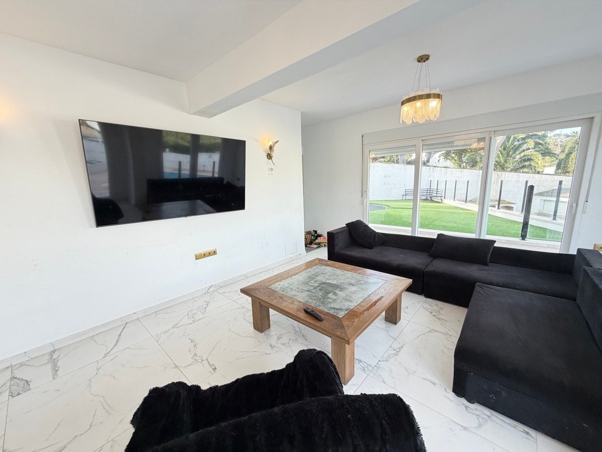 Modern living room with large sectional sofa, coffee table, and a wall-mounted TV, overlooking a green garden through large windows.