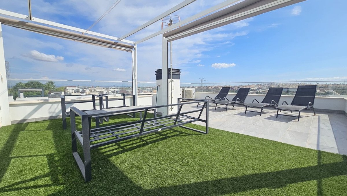 Spacious rooftop terrace with artificial grass, sun loungers, seating area, and panoramic city views under a blue sky.