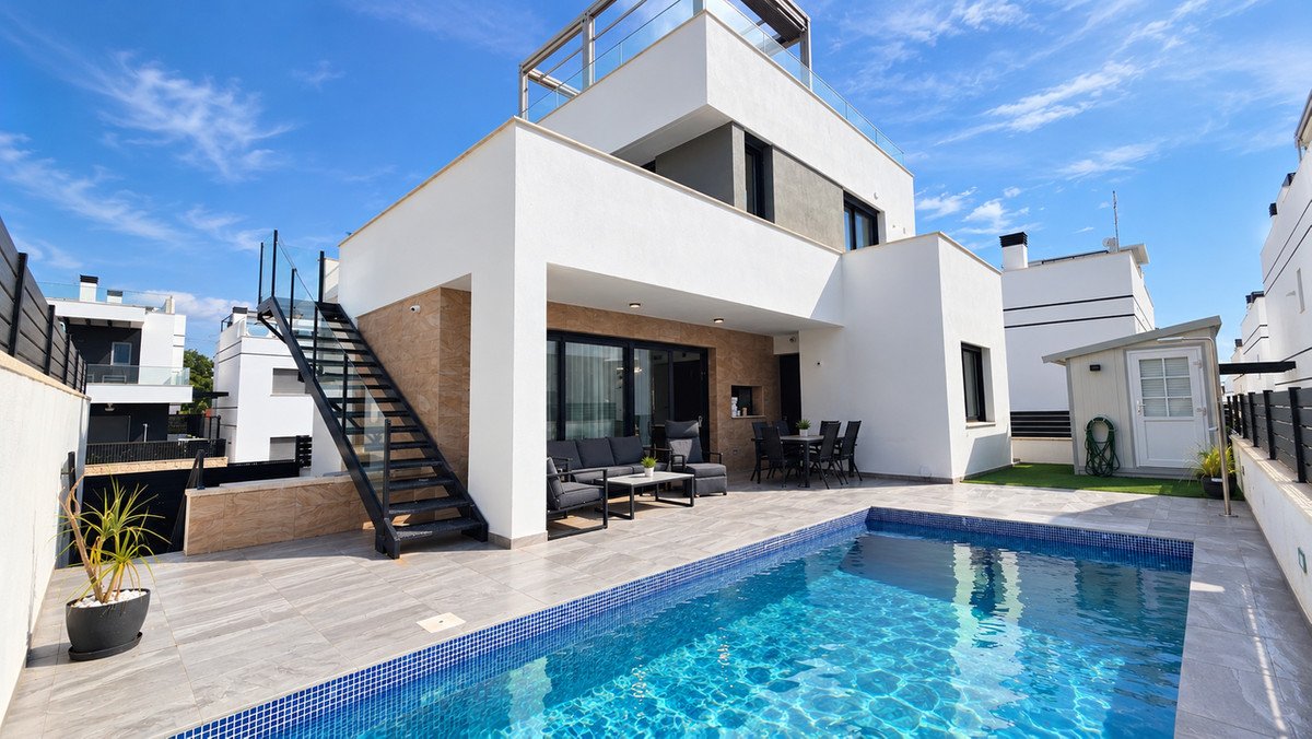 Modern villa exterior with a swimming pool, outdoor seating area, dining table, and rooftop terrace on a sunny day.