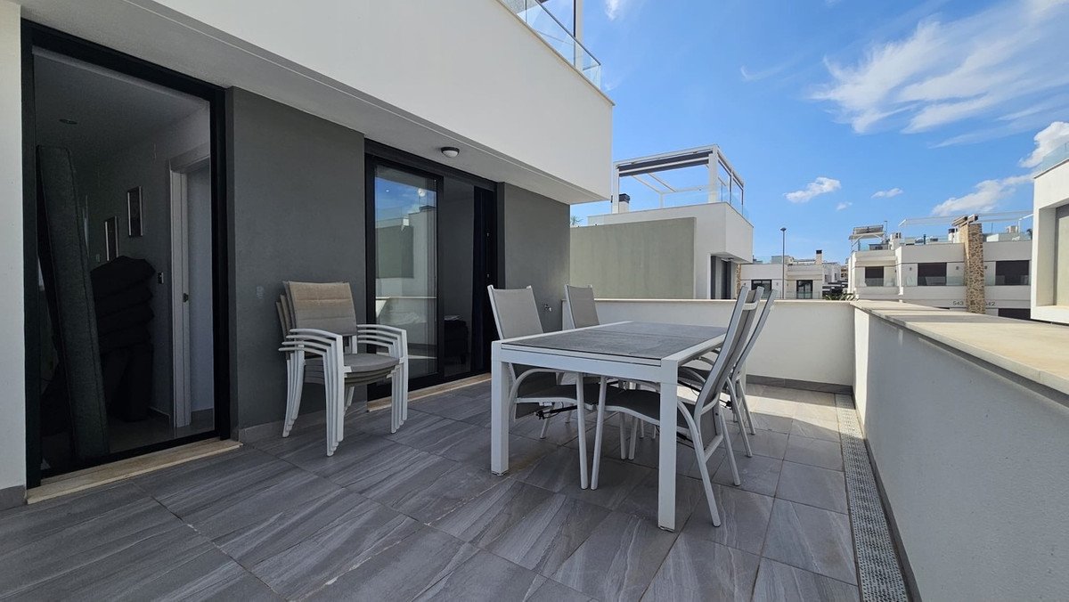 A terrace with a dining set and stacked chairs, featuring sliding doors leading indoors, overlooking other buildings under a blue sky.
