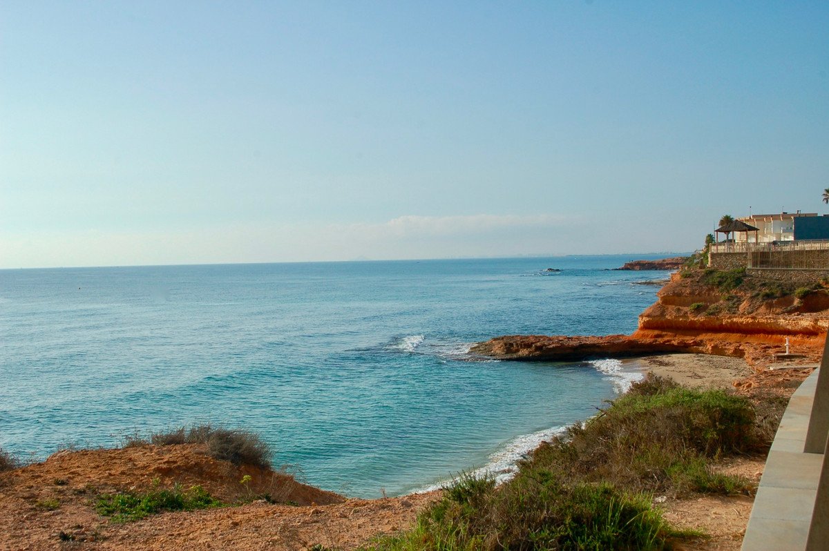 View of a coastal area with cliffs, a small beach, blue sea, and buildings on the clifftop.