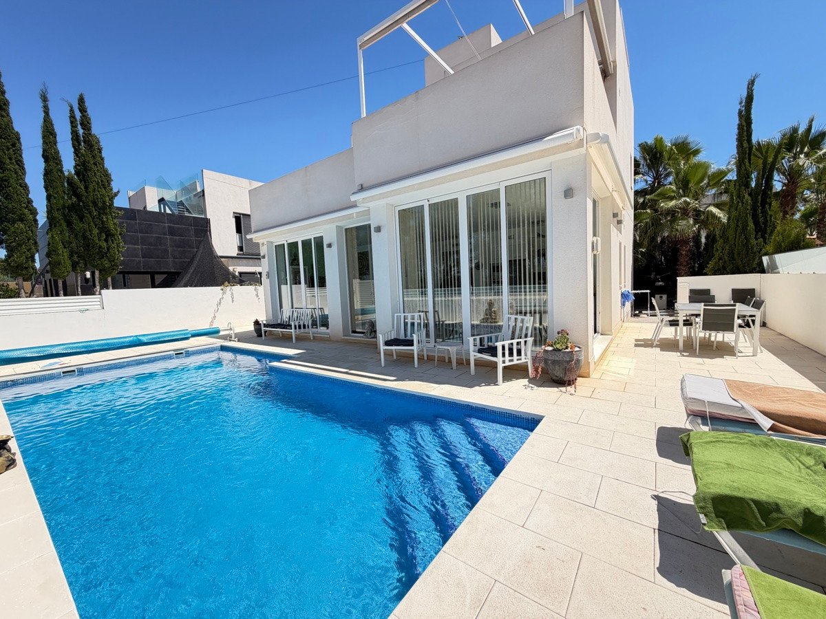 Modern villa exterior with a large blue swimming pool, tiled terrace, outdoor dining area, and lounge chairs under a sunny blue sky.