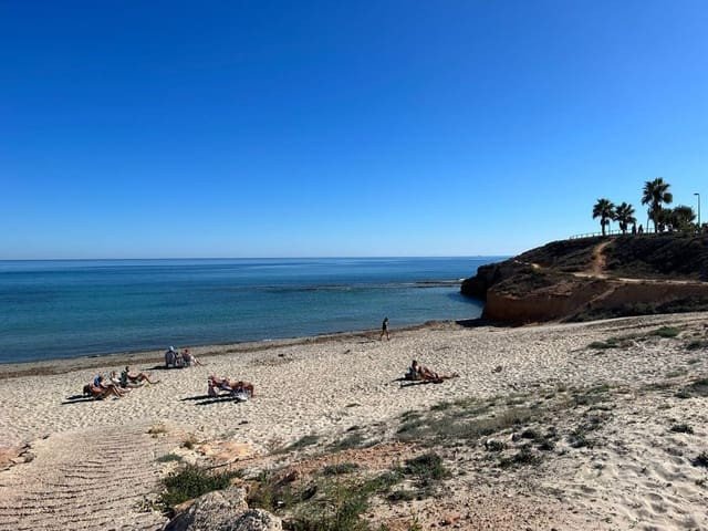 A wide sandy beach with people sunbathing, overlooking a calm blue sea with a cliff and palm trees on the right.