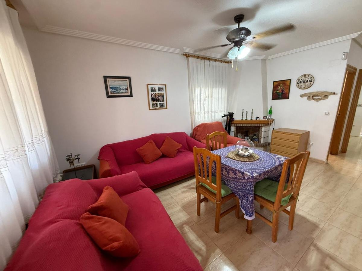 A cozy living and dining room with red sofas, a dining table, and a fireplace, featuring large windows with white curtains.