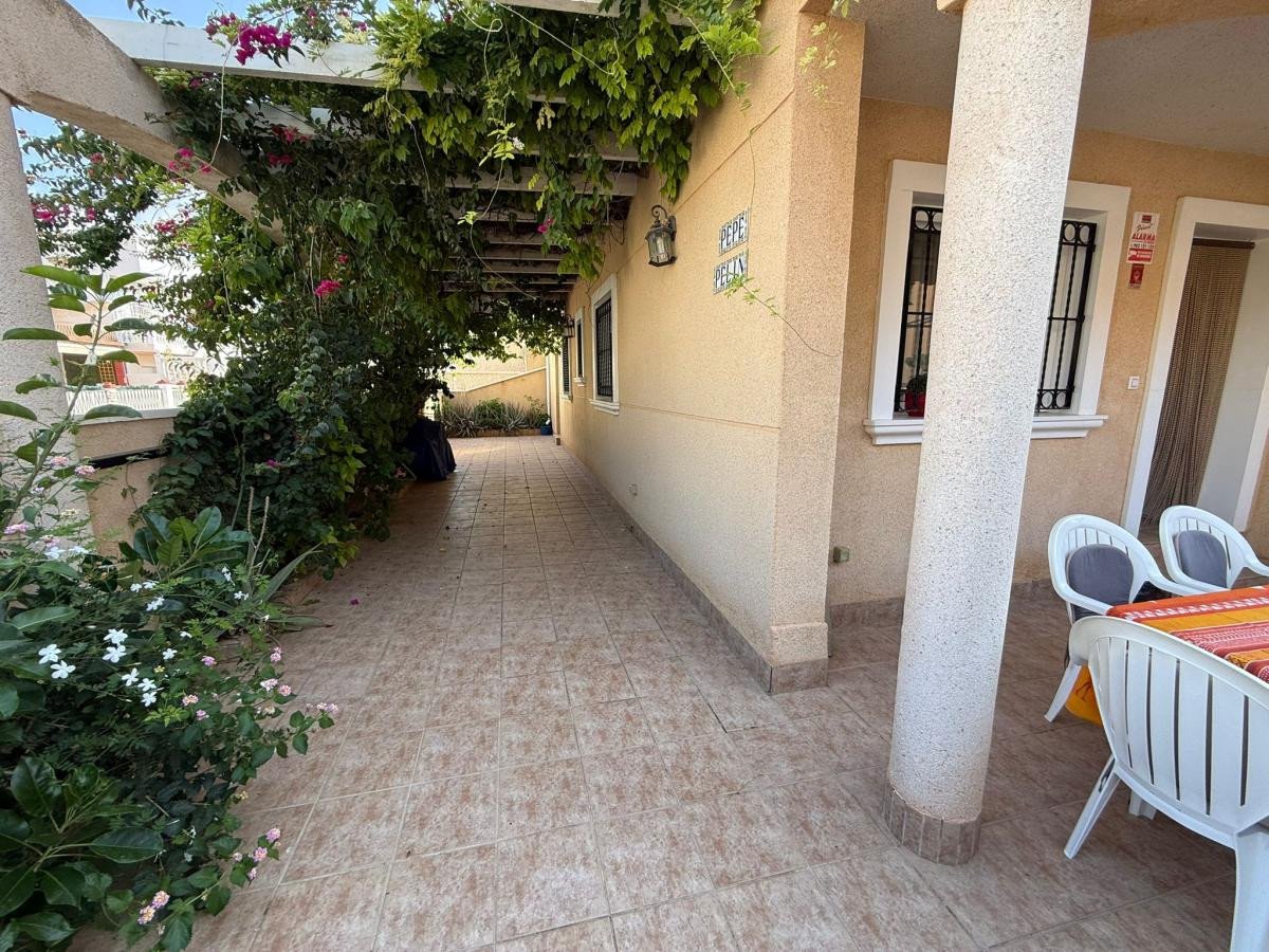 A covered terrace with a pergola entwined with vines and flowering plants, leading to a tiled patio with outdoor seating.