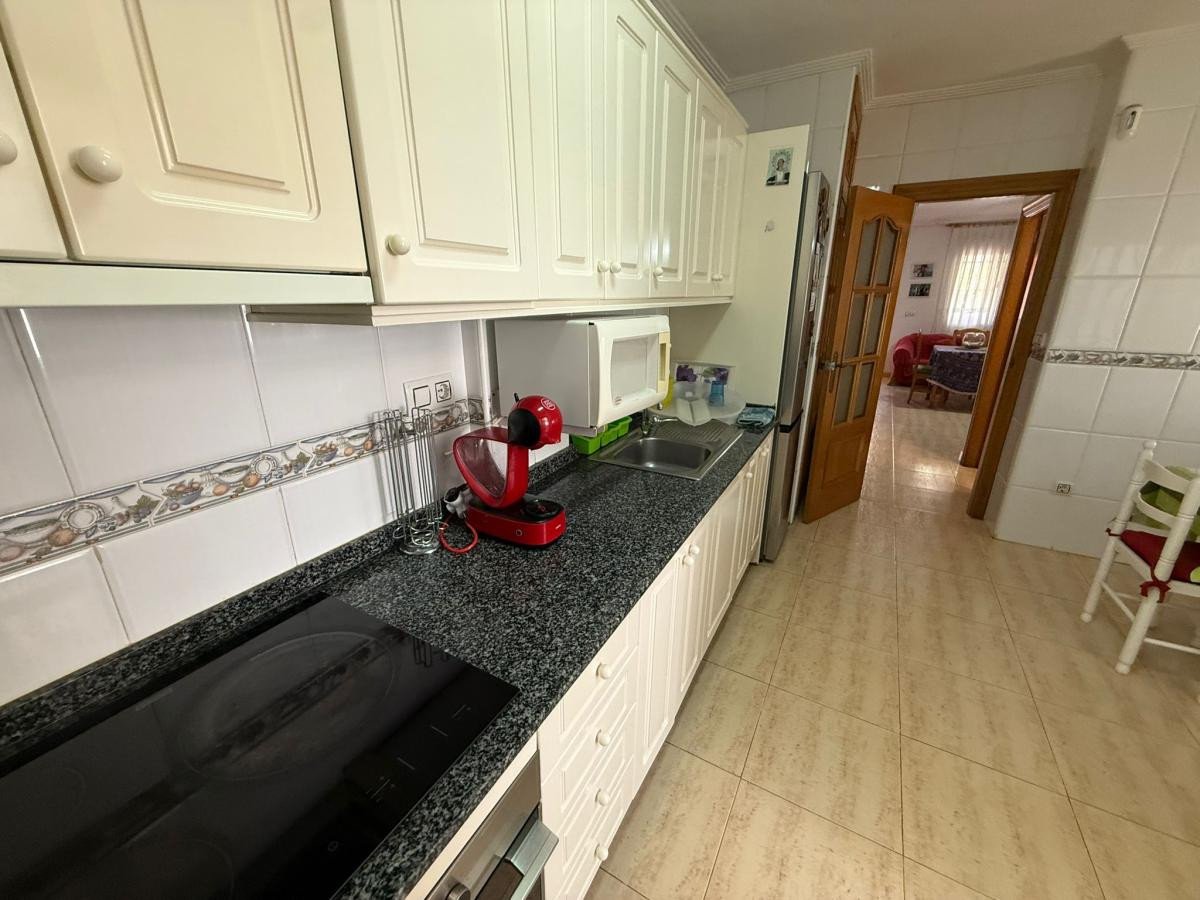 A modern kitchen features white cabinets, a granite countertop, an induction hob, and a red coffee machine. The doorway leads to a living room.