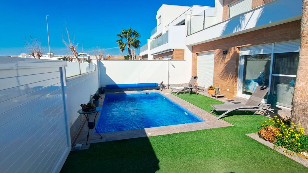 Modern villa backyard with a private swimming pool, sun loungers, and artificial grass, featuring a palm tree and contemporary architecture.