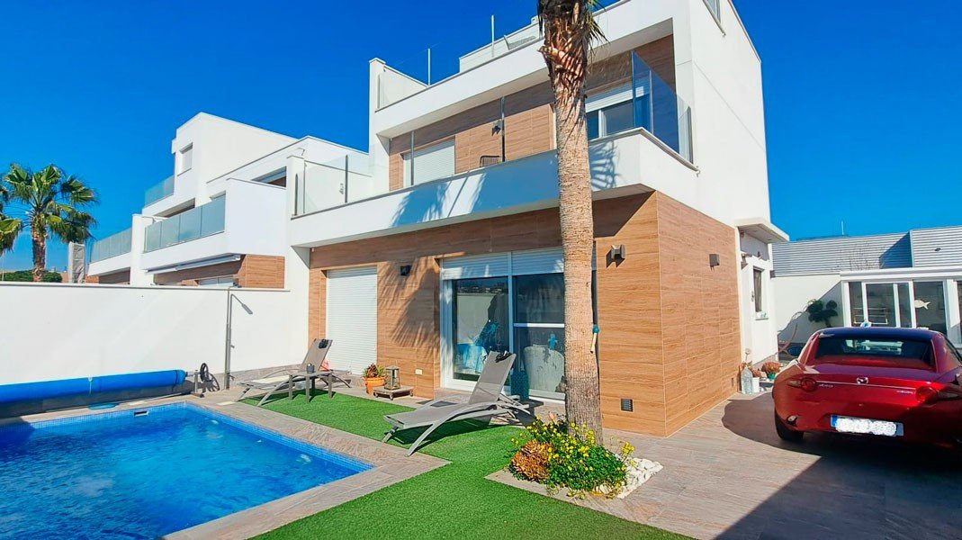 Modern villa with a private swimming pool, sun loungers, artificial grass, and a red car parked outside on a sunny day.