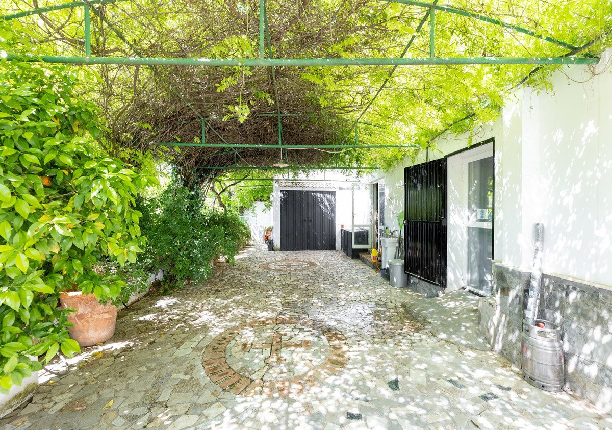 A covered driveway with a garage, lush greenery, and decorative stone paving under a vine-covered pergola.