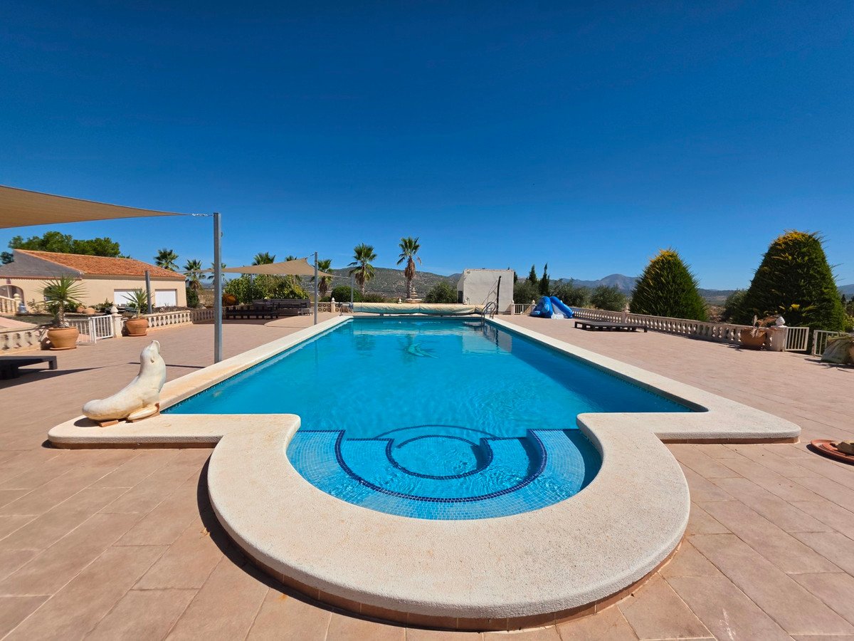 Large swimming pool with steps, a blue slide, outdoor seating area, and a mountain view under a clear blue sky.