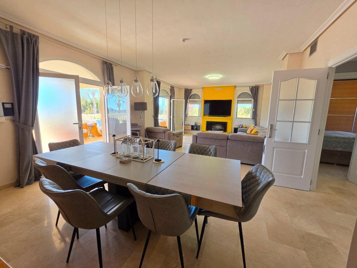 Modern dining area with plush chairs, leading to a living room with a fireplace and TV, with terrace access visible.