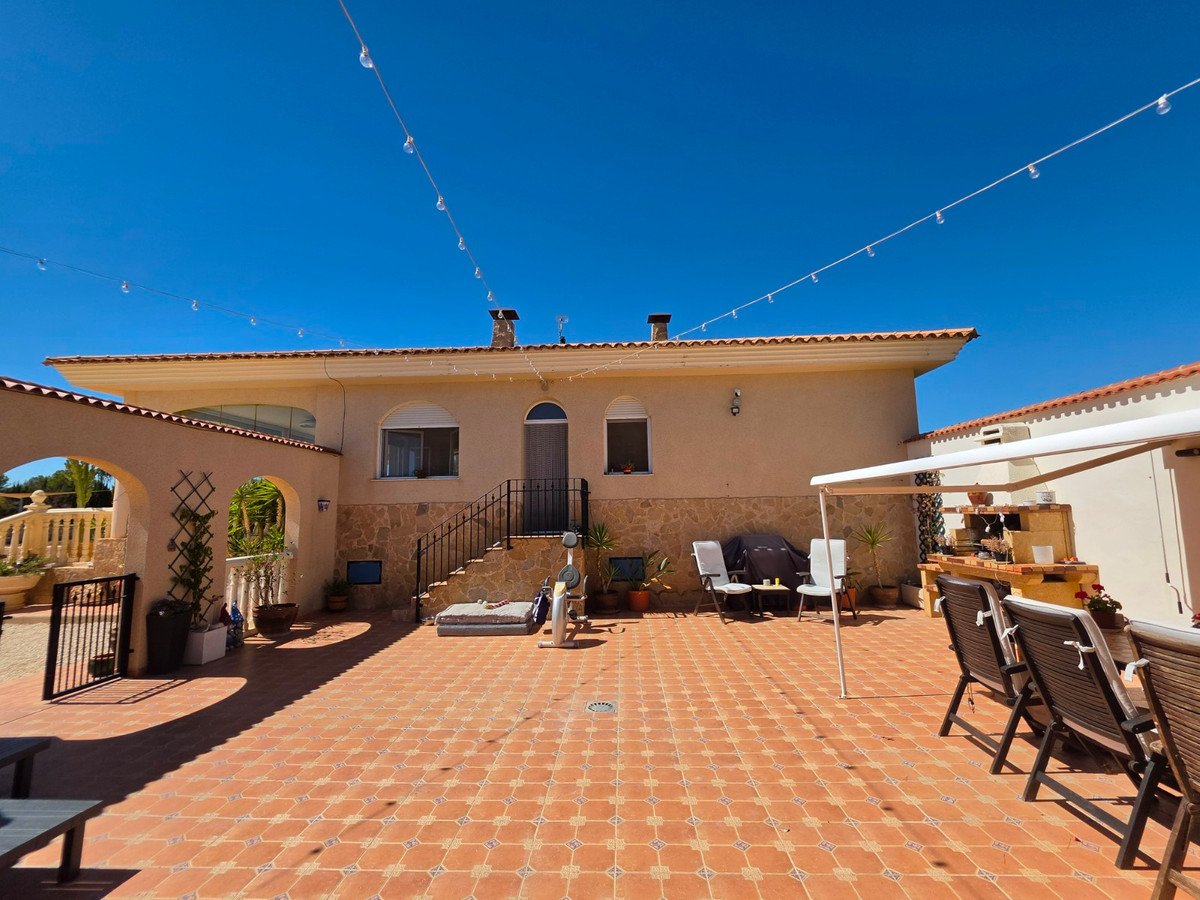 Tiled patio with outdoor kitchen, seating area, BBQ, and string lights under a clear blue sky.