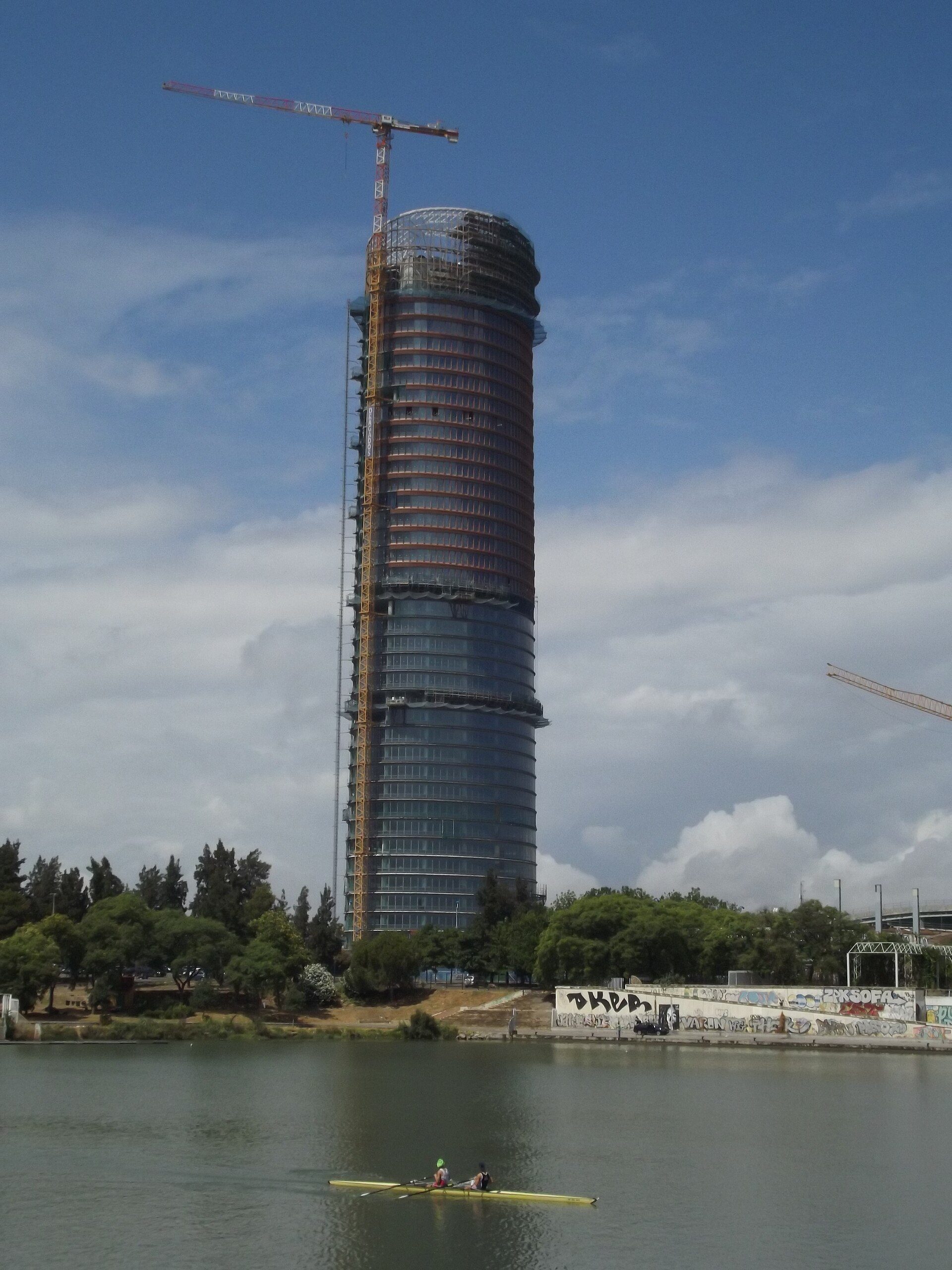 Aerial view of a modern high-rise building under construction with a crane, surrounded by trees and a body of water.