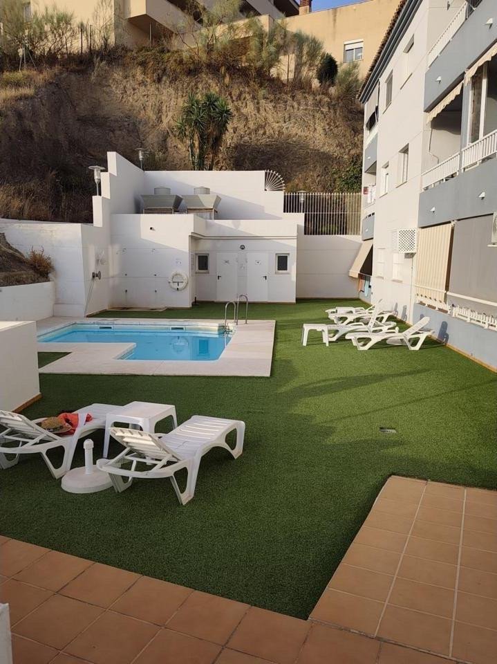 Outdoor pool area with artificial grass, sun loungers, and lounge chairs, next to a building and a hillside.