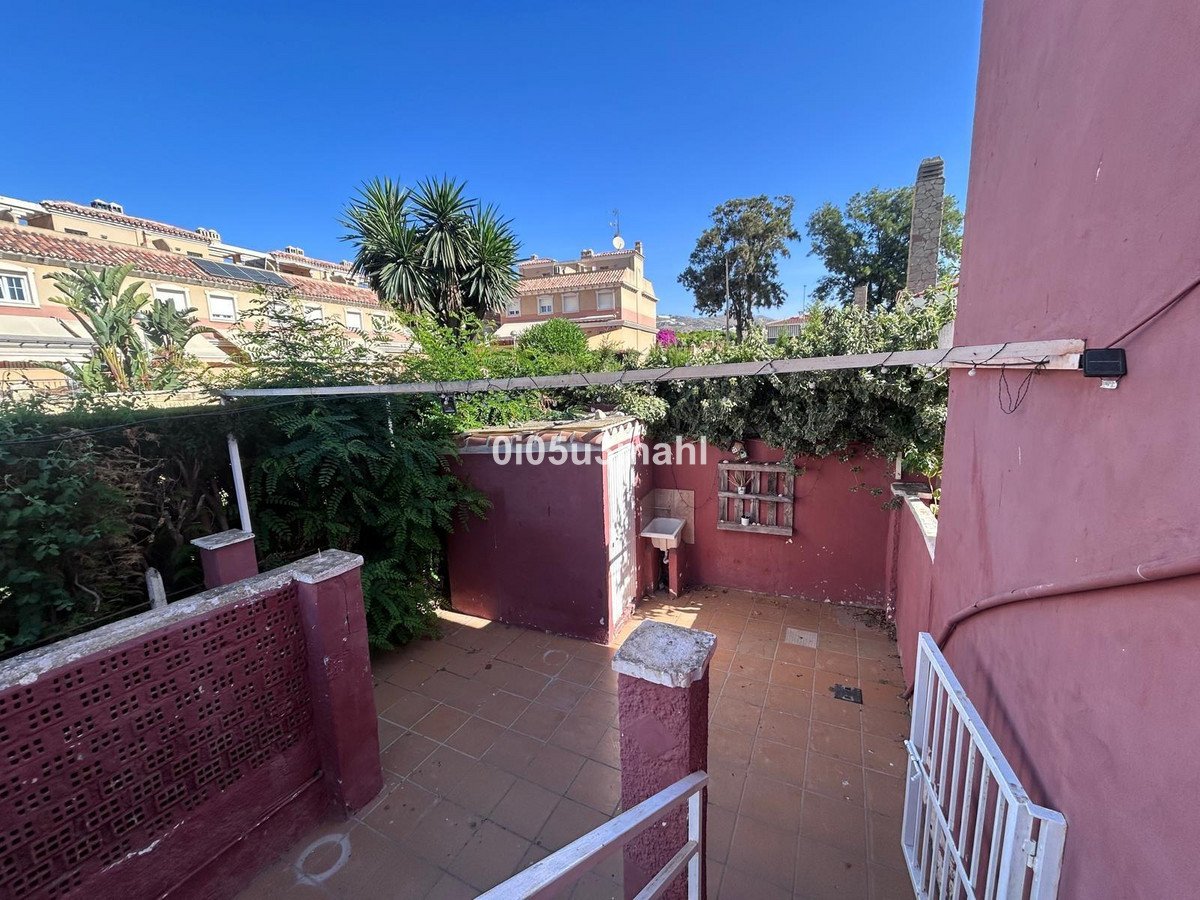 A small, pink-walled terrace with a sink, tiled floor, and lush green plants under a clear blue sky.