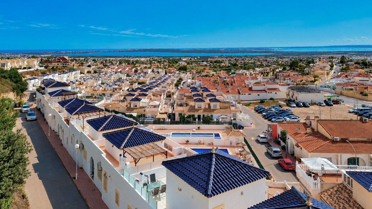 Aerial view of a townhouse complex with blue-tiled roofs, swimming pools, and a distant sea view under a clear blue sky.