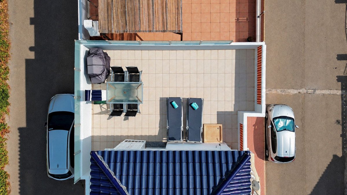 Aerial view of a sunny terrace with lounge chairs, a dining set, and two cars parked nearby.